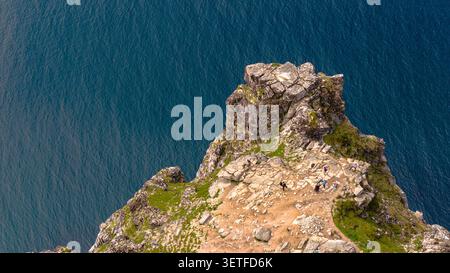 Aerial view of a dramatic rocky peak covered in moss and lichen, surrounded by deep teal ocean waters in Lofoten. Hikers explore the rugged Stock Photo