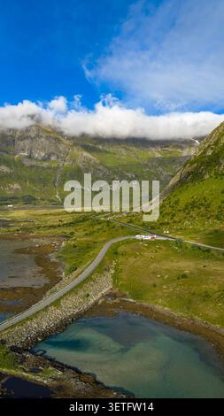 Scenic mountain valley in Lofoten featuring turquoise alpine lake, winding road through lush green slopes, dramatic peaks with cloud cover Stock Photo