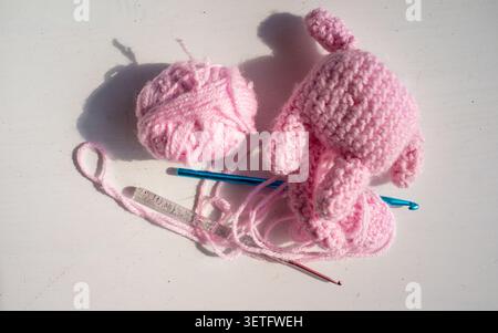 Pink knitted baby booties beside yarn ball, crochet hook, loose thread on white surface with soft shadow, handmade craft setup, cozy pastel textile st Stock Photo