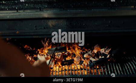 Barbecue grill glows with burning charcoal. Flames dance on hot coals. Focused hand adjusts cooking food. Heat radiates from the fire. Perfect for out Stock Photo