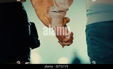 Close-up of two people holding hands. One wears white shirt sleeves. The other's dark clothing is visible. Soft focus background suggests evening ligh Stock Photo