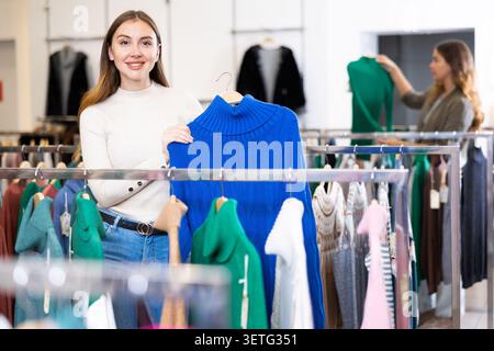 Positive casual female holding blue pullover on a hanger while choosing clothes in a store Stock Photo