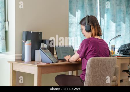 A dedicated student engages in online study using multiple devices in a comfortable workspace at home. Stock Photo