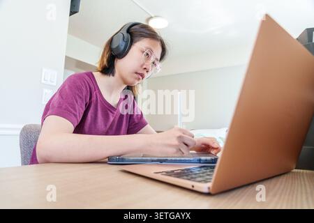 A young woman studies online using a laptop and digital tablet immersed in her work in a contemporary setting. Stock Photo