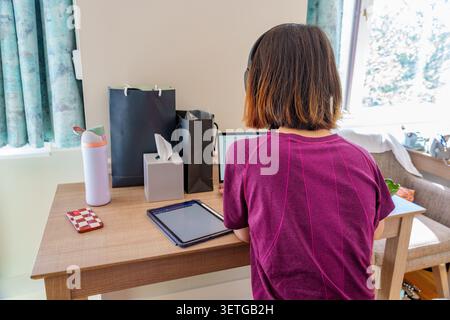 Back view of a young female student studies at a home workspace utilizing technology and surrounded by personal items showcasing a cozy learning envir Stock Photo