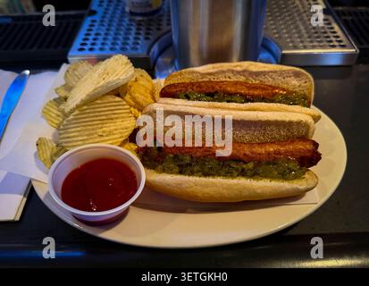 Two hot dogs topped with bright green relish are served in soft buns alongside ridged potato chips and a cup of ketchup, arranged on a plate at a casu Stock Photo