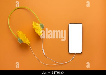 Creative composition of smartphone mockup with blank screen connected to flower headphones by cable on orange background. Minimal mobile app presentat Stock Photo
