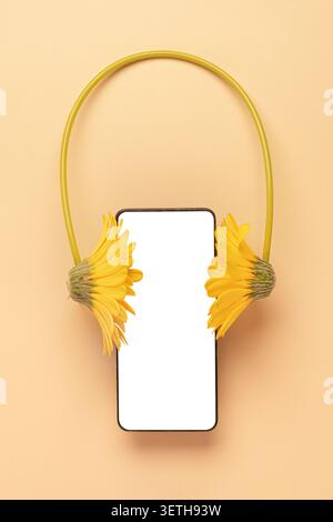 Creative composition of smartphone mockup with yellow flower headphones on soft beige background. Concept of music app, podcast and audiobook streamin Stock Photo