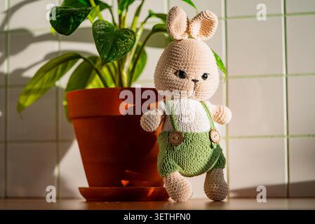 Crochet bunny amigurumi doll on a wooden table next to a terracotta potted houseplant, bathed in warm sunlight for a cozy, handmade home decor scene Stock Photo