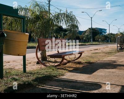 Wooden bench with curved legs sits near a small palm tree. Yellow bucket rests by a green structure. Sunlight creates long shadows on dry ground. Stre Stock Photo