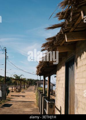 Sunlit path winds through a quiet village. Light-colored brick building with thatched roof lines the street. Clear blue sky stretches overhead with di Stock Photo