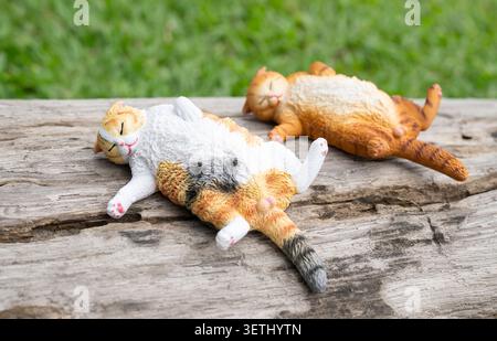 Chiang Mai, Thailand - 19 June 2026 : Calico and orange tabby cat figurines lying on their backs on old weathered log with green nature background Stock Photo