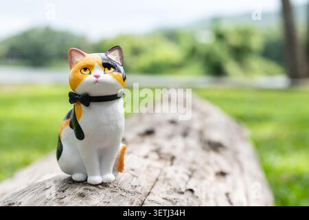 Chiang Mai, Thailand - 19 June 2026 : Calico cat figurine with black bow tie sitting on old weathered log with unimpressed expression and green mounta Stock Photo