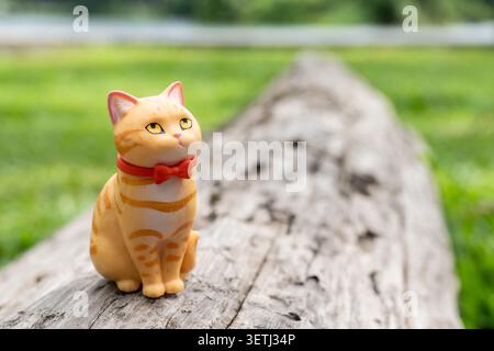 Chiang Mai, Thailand - 19 June 2026 : Orange tabby cat figurine with red bow tie sitting on old weathered log with unimpressed expression and nature b Stock Photo
