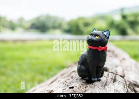 Chiang Mai, Thailand - 19 June 2026 : Black cat figurine with red bow tie sitting on old weathered log with unimpressed expression and green nature ba Stock Photo