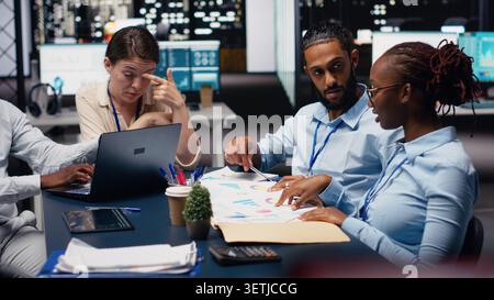 Multiethnic team working together in office, looking over paperwork on laptop. Diverse coworkers collaborating on strategy planning, preparing company data insights before deadline. Stock Photo