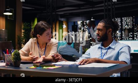 Middle Eastern man in wheelchair and woman CEO collaborating at night in corporate office, working on project with documents. Diverse colleagues analyze research data, disability-friendly workspace. Stock Photo