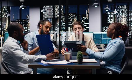 Diverse team of employees in office looking over paperwork documents late at night. Coworkers at work cooperating, analyzing company budget reports documentation files before deadline Stock Photo