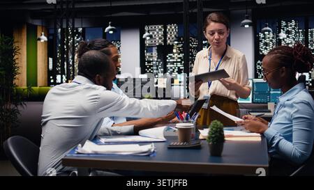 Evaluating staff performance, tablet and manager gathering employees in office, offering task completion feedback. Team leader reviewing KPIs, overseeing work done by multiethnic staff. Stock Photo
