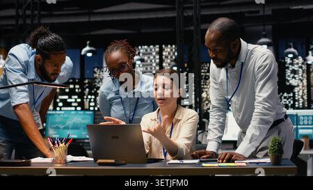Corporate team gathered around office desk collaborating on project and sharing business intelligence insights. Group working late into the night, focusing on forecasting and task management. Stock Photo