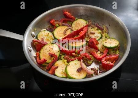 Zucchini, bell peppers, onions, and celery are sauteed in a stainless steel pan on a black stove top, cooking a healthy vegetarian meal made from vege Stock Photo