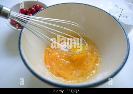 Eggs are beaten with a whisk in a bowl, preparation for a cherry cake. Seasonal baking with homegrown fruits, selected focus, narrow depth of field Stock Photo