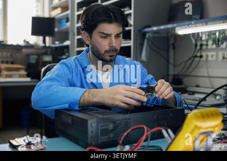A technician man soldering in a distinctive blue lab coat is intently repairing a computer Stock Photo