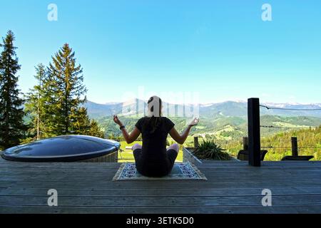 Woman meditating on terrace overlooking mountains. Woman sits cross-legged on a wooden terrace meditating toward a wide mountain valley, with Stock Photo