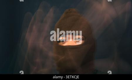 A male wearing a brown balaclava looks steadily forward as blurred ghostly hands hover in heavy motion around his face and head area. The haunting and Stock Photo