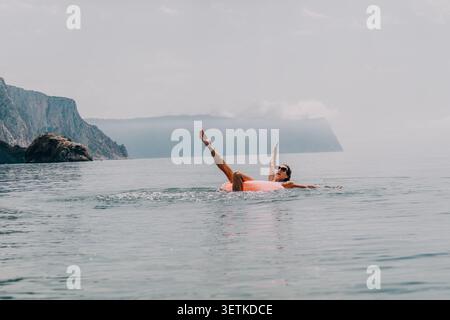 Woman inflatable ocean joyful woman floats on an orange ring in calm sea water against scenic coastal cliffs and mountains Stock Photo