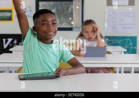 Diverse child classmates raising hand and using tablets in classroom at white desks. Edtech, devices, students, learning, focus, window, yellow Stock Photo