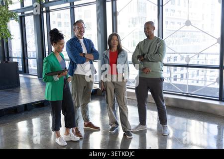 Diverse coworkers in casual clothes standing near metal windows in office holding tablet and phone. Openplan, glazing, urban, daylight, collaboration, platform, plants Stock Photo