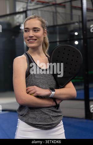 Female standing on padel court holding racket and ball, wearing grey tank and white skirt. Woman, athlete, racquet, activewear, ponytail, smartwatch, glass Stock Photo