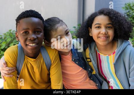 Diverse pre-teen friends sitting on bench in courtyard with backpacks, denim jacket, gray hoodie. Students, outdoor, schoolyard, colorful, friendship, casual, daylight Stock Photo