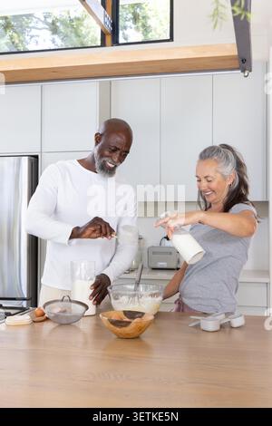 Couple making batter at home, senior African-American man scooping flour, woman pouring milk. Kitchen, baking, cooking, modern, island, countertop, ingredients Stock Photo