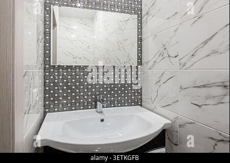 A modern bathroom featuring white marble-patterned tiles, a mosaic tile accent wall, a white basin sink, and a framed mirror above the sink. Bright an Stock Photo