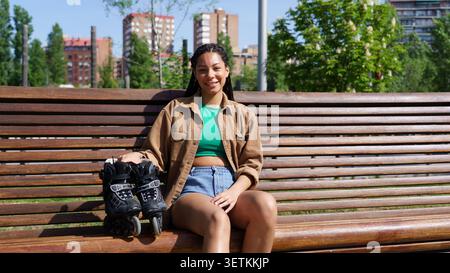 Smiling young woman with braids sitting on a wooden bench holding her inline skates in an urban park Stock Photo