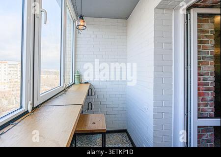 A cozy balcony with white brick walls, a wooden countertop by the window, and a hanging light Stock Photo