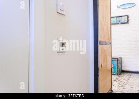 A bright interior shot focusing on a wall with a light switch and a thermostat. Partially visible is a wooden panel and a glimpse into an adjacent roo Stock Photo