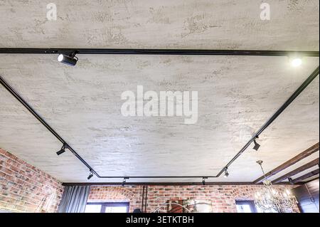 Looking up at a textured ceiling with black track lighting and spotlights. The room has exposed brick walls and a chandelier Stock Photo