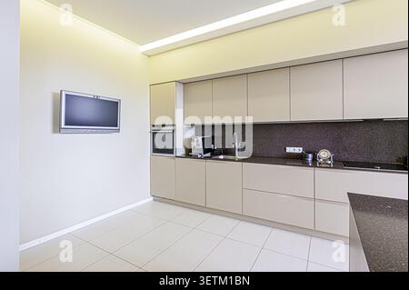 A modern kitchen with sleek beige cabinets, dark countertops, and a TV on the wall. The floor is light-colored tile Stock Photo