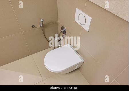 A modern, clean bathroom corner features a white toilet with a rounded lid. Beige tiles cover the walls and floor. A bidet, water valve, and toilet br Stock Photo