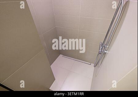 A clean shower stall with light beige tiles and white flooring, featuring a sleek metal shower fixture and a linear drain. The design is modern and mi Stock Photo
