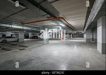 A well-lit, spacious underground parking garage with concrete floor and ceiling, visible pipes, support columns, and parked cars Stock Photo