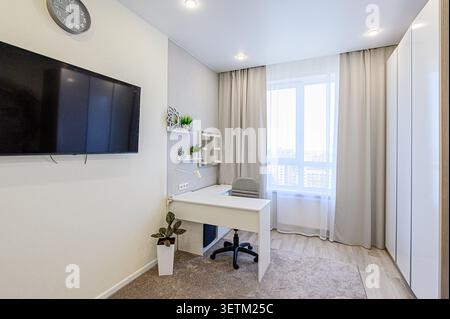 A modern room features a white desk with a gray chair near a window with curtains. A TV hangs on the wall, and there are shelves with decorative items Stock Photo