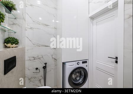 Modern bathroom with white marble-like tiles, a white washing machine neatly fitted into a built-in cabinet, and a white door with black hardware Stock Photo