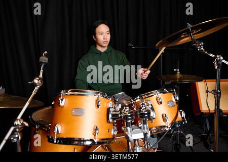 A young Asian man in casual clothes plays drums, focused in a recording studio. Stock Photo