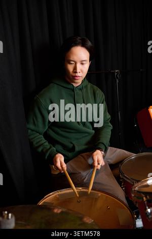 Casual and focused, a young Asian man plays the drums in a vibrant recording studio. Stock Photo