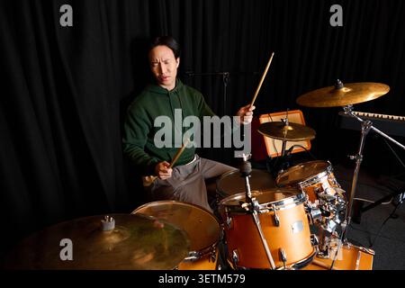 A handsome young Asian man passionately plays drums in a stylish recording studio. Stock Photo