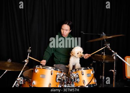 A young man plays drums passionately while his fluffy dog sits next to him. Stock Photo
